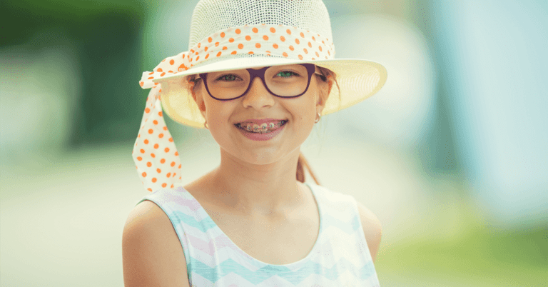 A girl in a white sun hat with an orange polka-dot ribbon, glasses and braces from Levine Orthodontics in Fort Myers, Cape Coral or Lehigh Acres, FL, smiles outdoors.