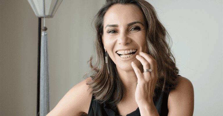 Showcasing the confidence that comes from adult orthodontic treatment at Levine Orthodontics in Fort Myers, Cape Coral or Lehigh Acres, FL, a woman with shoulder-length brown hair in a sleeveless black top smiles and touches her face indoors beside a lamp.