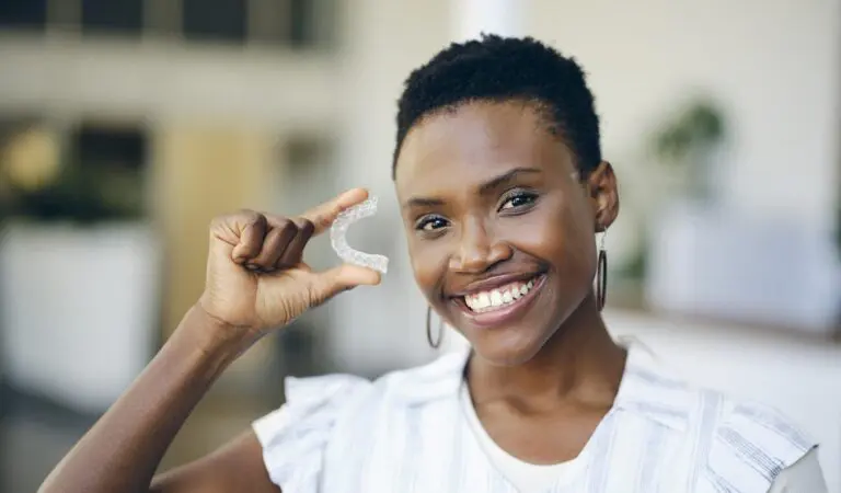 A woman smiles and holds up a clear dental aligner with her right hand in a bright indoor setting, showcasing the benefits of Levine Orthodontics in Fort Myers, Cape Coral or Lehigh Acres, FL.