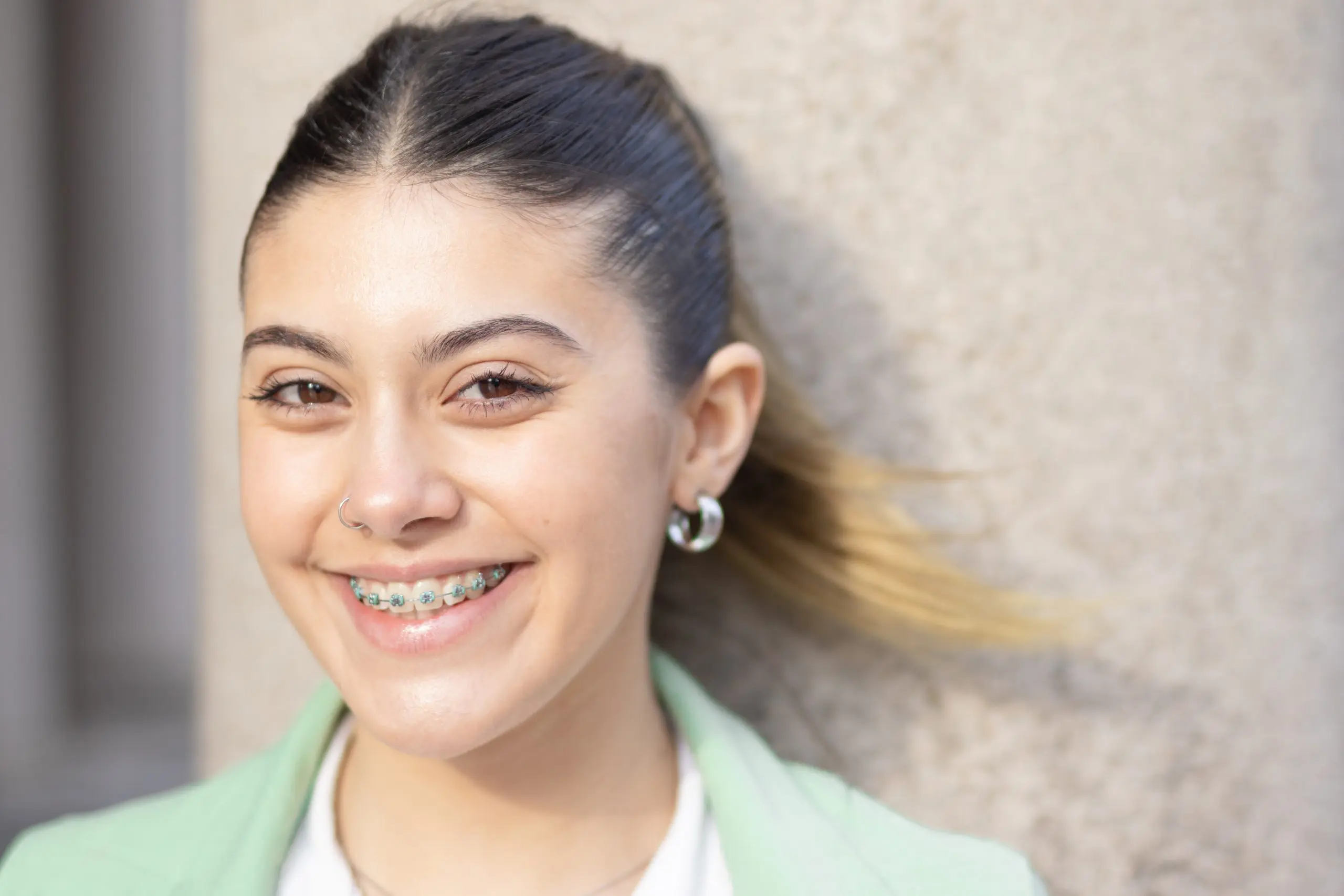 At Levine Orthodontics in Lehigh Acres, FL, a young woman with long hair in a ponytail smiles confidently at the camera, showing her braces. She wears hoop earrings and a light green jacket while standing against a stone wall.