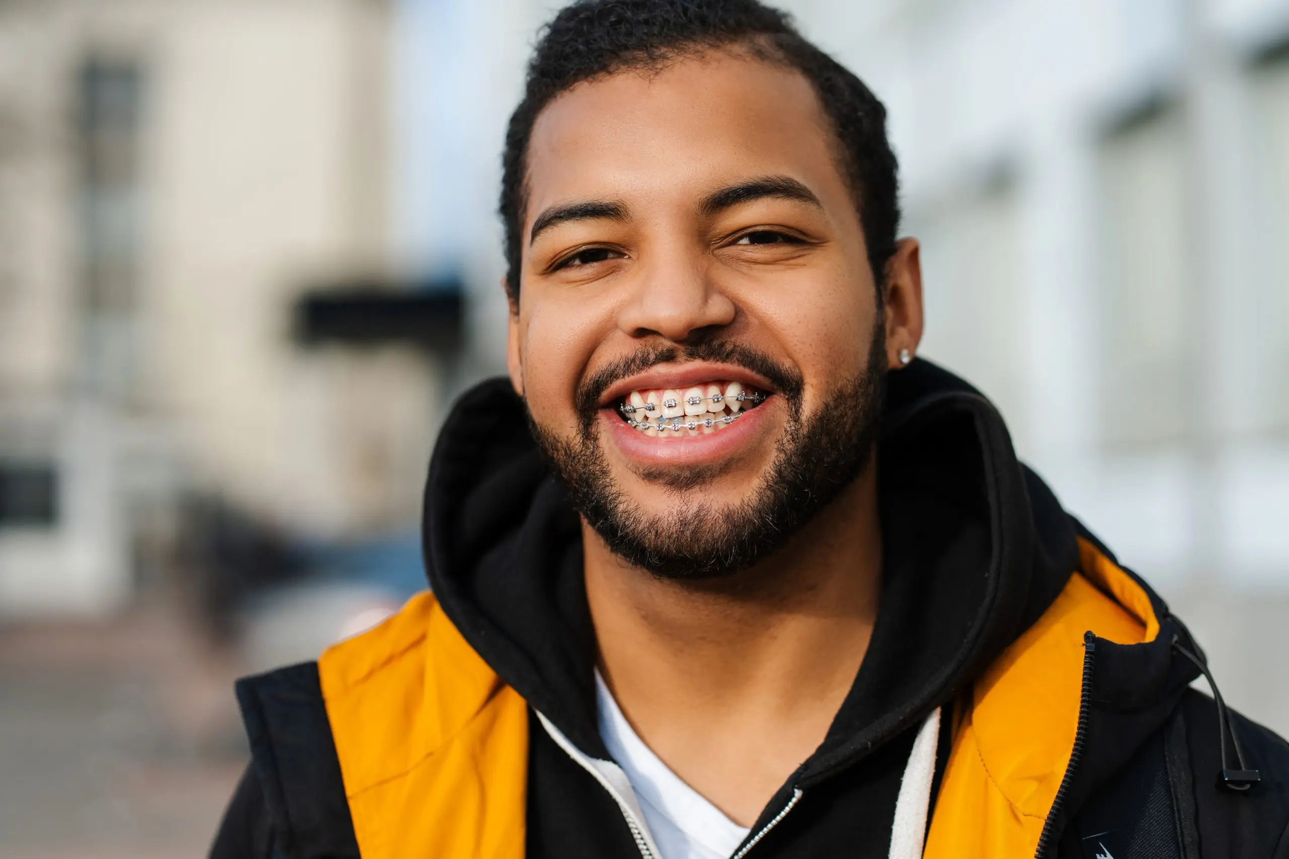 A bearded young man with an underbite, wearing braces, a black hoodie, and a yellow jacket, smiles outdoors an example of the confident results achieved at Levine Orthodontics in Fort Myers, Cape Coral or Lehigh Acres, FL.