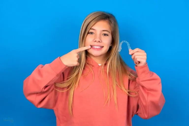 A girl in a pink hoodie points to her teeth and holds a clear aligner, highlighting alternatives to braces available at Levine Orthodontics in Fort Myers, Cape Coral or Lehigh Acres, FL, against a bright blue background.