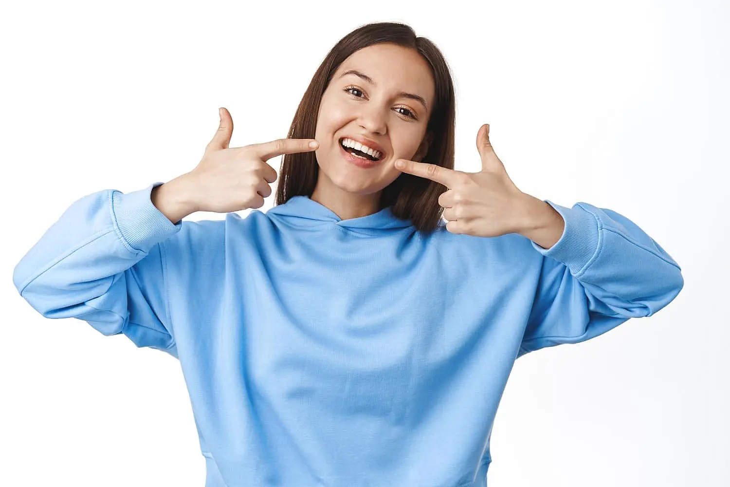 A woman in a light blue hoodie smiles and points to her metal braces with both index fingers, proudly displaying her results from Levine Orthodontics in Fort Myers, Cape Coral or Lehigh Acres, FL.