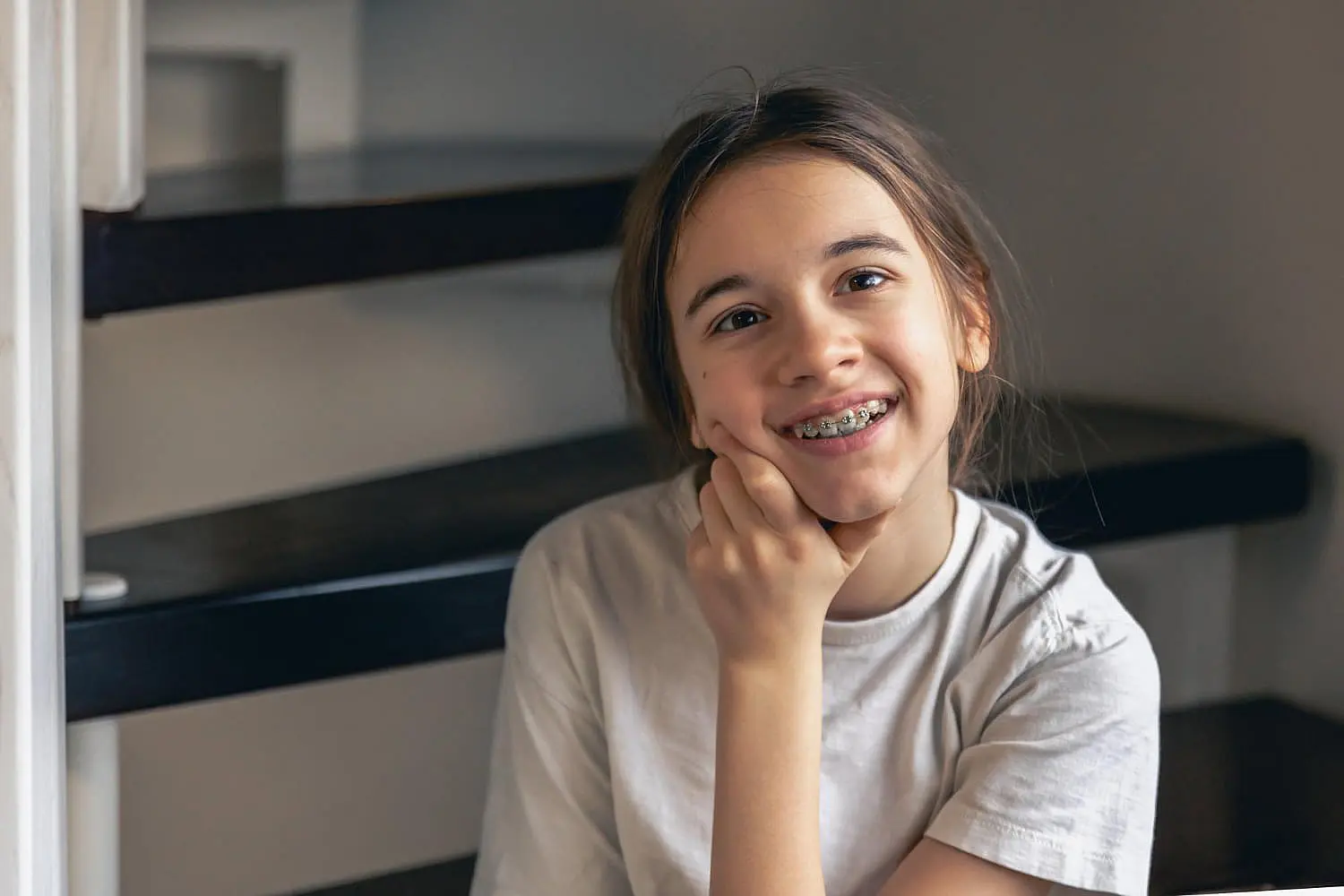 A young person with long brown hair and braces, representing the benefits of adult orthodontics at Levine Orthodontics in Fort Myers, Cape Coral or Lehigh Acres, FL, sits indoors on dark wooden stairs, resting their chin on their hand with a slight smile.