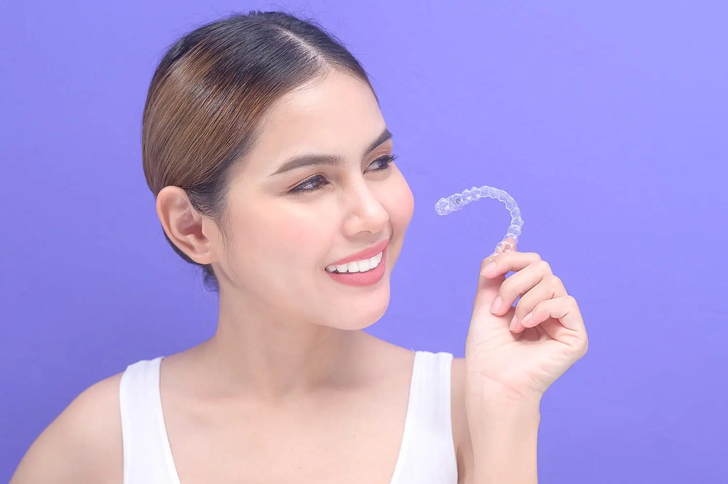 A smiling woman holds a clear dental aligner against a purple background, contemplating the flexible Invisalign payment plans at Levine Orthodontics in Fort Myers, Cape Coral or Lehigh Acres, FL.