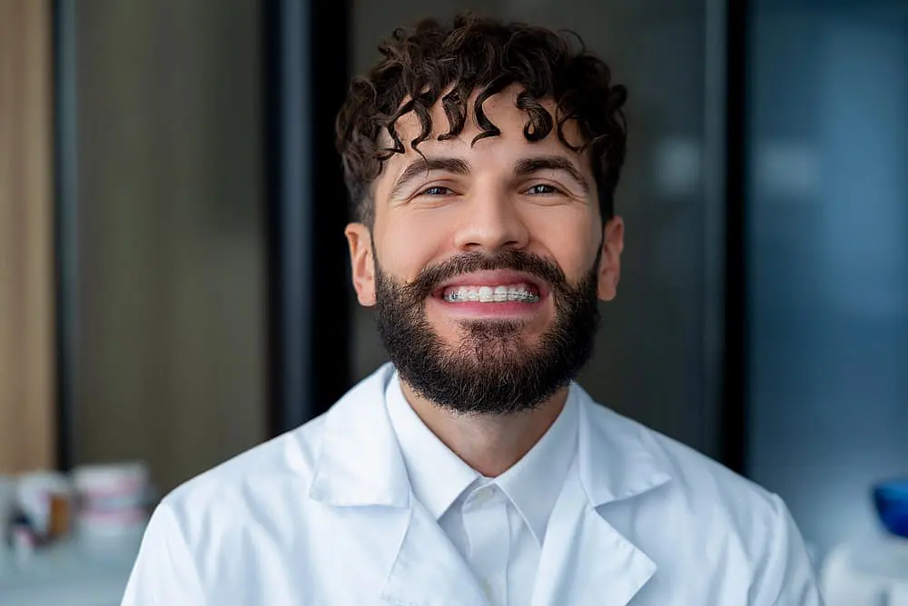In an indoor setting, a bearded individual with curly hair confidently smiles in a white lab coat, with bright lights emphasizing the clear braces. Visit Levine Orthodontics in Cape Coral, FL.