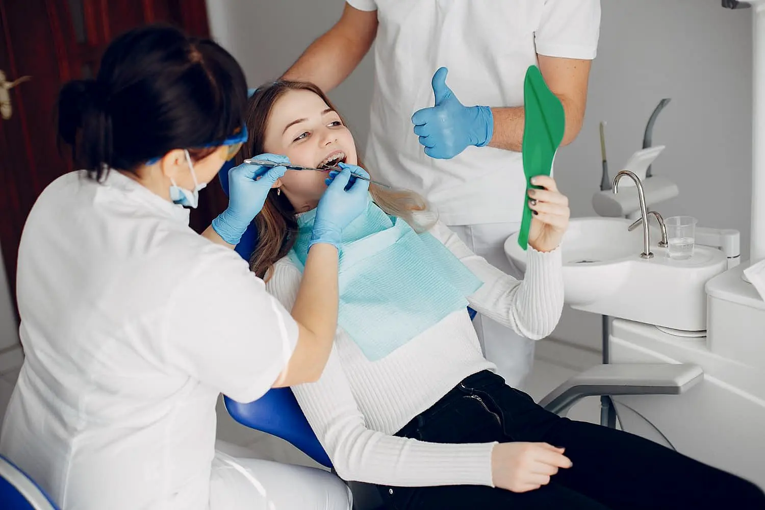 In a well-lit dental office, a young woman in the chair watches as her braces are adjusted to correct an overbite, holding a green mirror. Nearby stands an assistant at Levine Orthodontics in Fort Myers, Cape Coral or Lehigh Acres, FL.