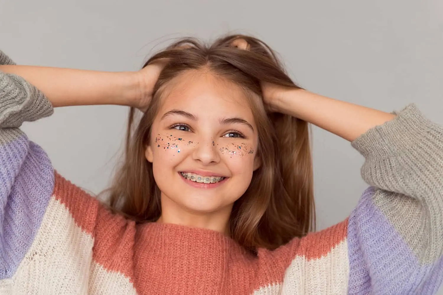 A smiling girl with traditional braces and glittery cheeks, dressed in a striped sweater, confidently holds her hair back against a plain background—showcasing the positive results achieved at Levine Orthodontics in Fort Myers, Cape Coral or Lehigh Acres, FL.