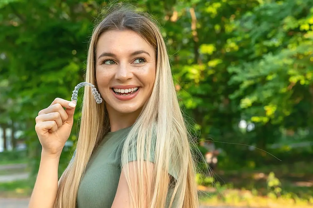 A smiling woman with long blonde hair holds Clear Aligners outdoors amid green trees, showcasing Levine Orthodontics in Fort Myers, Cape Coral or Lehigh Acres, FL.