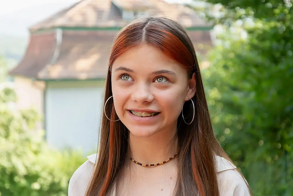 A teen girl with long brown hair, hoop earrings, and metal braces treated at Levine Orthodontics in Fort Myers, Cape Coral or Lehigh Acres, FL smiles outdoors amid greenery and a building.