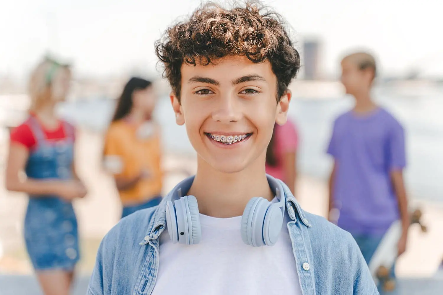 A smiling teenage boy with curly hair and metal braces, wearing headphones around his neck, stands outdoors with friends in the background—an example of the confident smiles created by Levine Orthodontics in Fort Myers, Cape Coral or Lehigh Acres, FL.