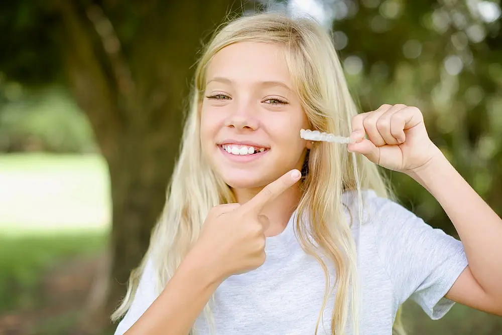 A smiling girl with long blonde hair stands outdoors, holding and pointing to a clear aligner, highlighting the benefits of visiting Levine Orthodontics in Fort Myers, Cape Coral or Lehigh Acres, FL for a children's orthodontist.
