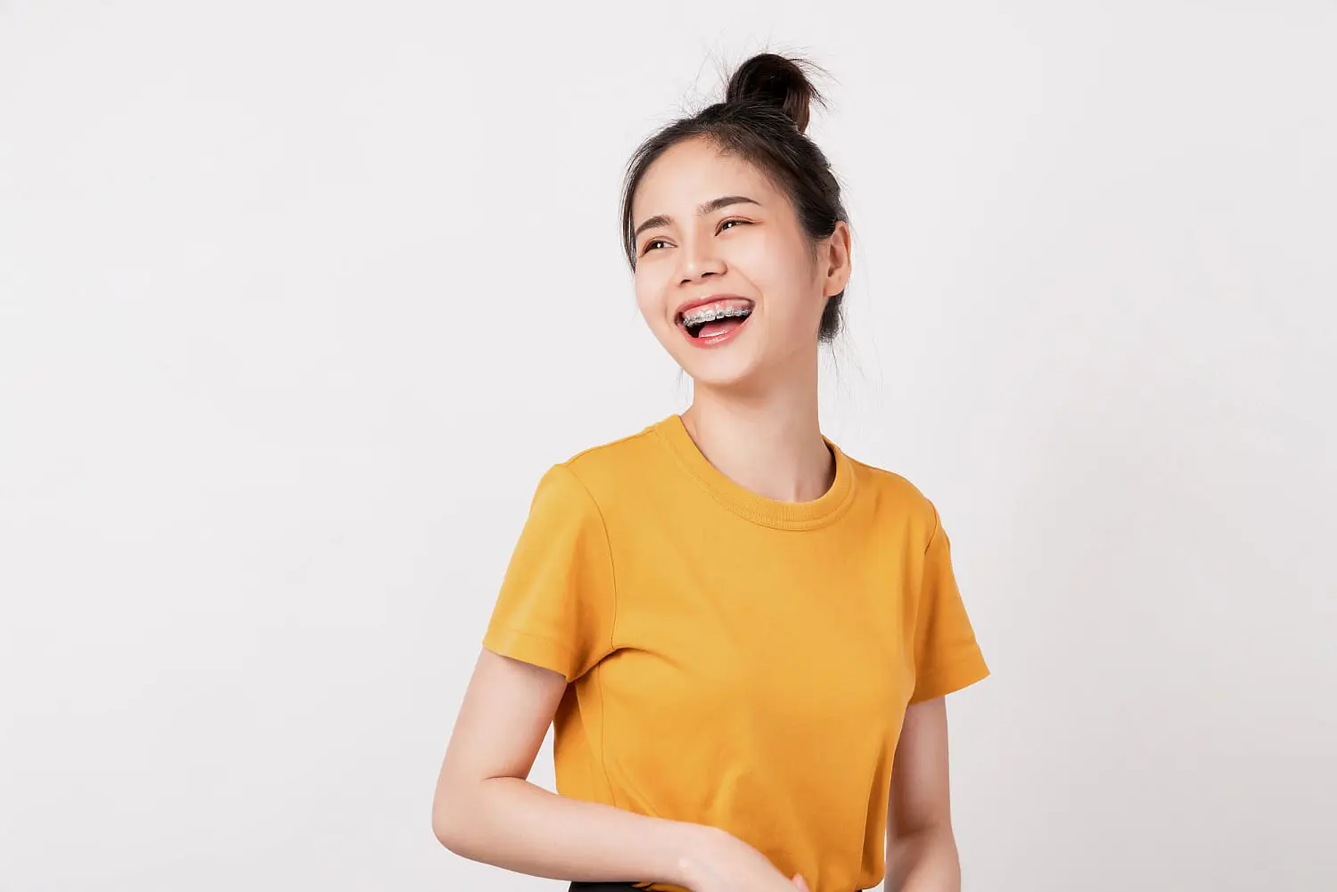 A young woman with metal braces, wearing a yellow t-shirt, smiles and looks to the side against a plain white background, representing Levine Orthodontics in Cape Coral, FL.
