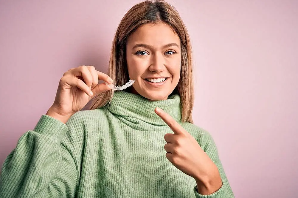 A woman in a green sweater smiles and points at Invisalign® Clear Aligners she's holding, standing against a light pink background—a service offered by Levine Orthodontics in Fort Myers, Cape Coral or Lehigh Acres, FL.