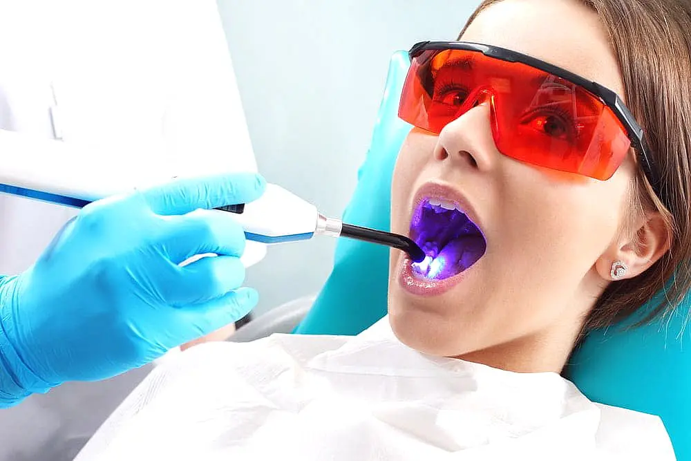 A person at Levine Orthodontics in Fort Myers, Cape Coral or Lehigh Acres, FL receives dental treatment, comparing Invisalign vs braces, while using a blue light device with protective red glasses and a dental bib.