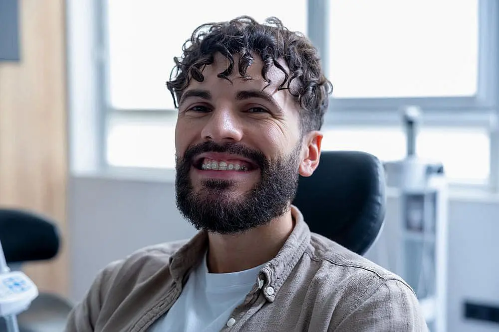 Sitting by a window in a neutral-colored shirt, a curly-haired, bearded man smiles with ceramic braces from Levine Orthodontics in Fort Myers, Cape Coral or Lehigh Acres, FL.