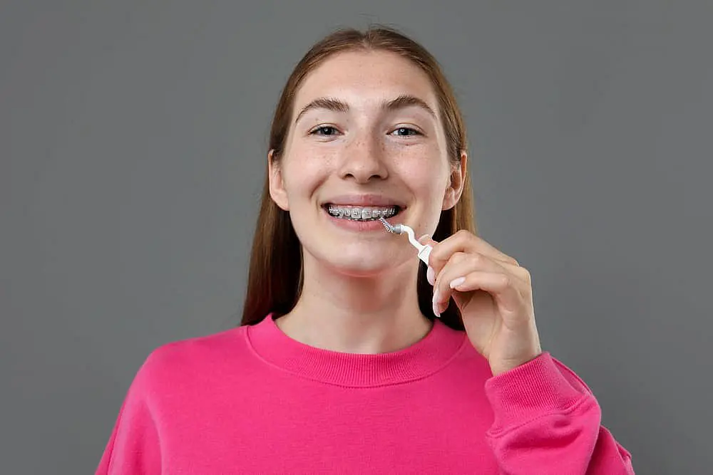 In Fort Myers, Cape Coral or Lehigh Acres, FL, a young person with long hair and metal braces at Levine Orthodontics uses a dental floss pick while wearing a pink shirt against a gray background.