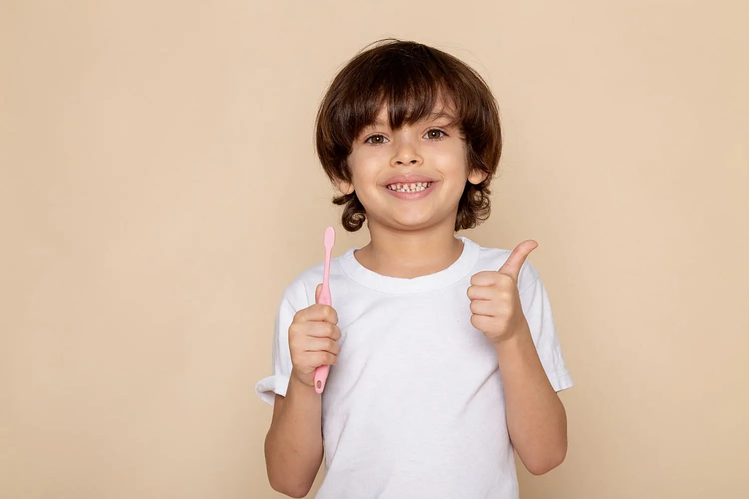 A young child in a white t-shirt, with braces and a bright smile, holds a pink toothbrush and gives a thumbs-up after visiting Levine Orthodontics in Fort Myers, Cape Coral or Lehigh Acres, FL. The child stands against a plain beige background.