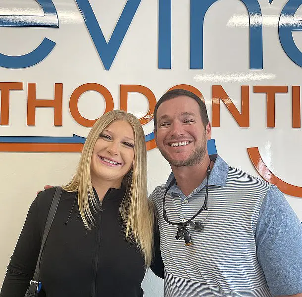Two people smile together in front of a wall sign displaying "ORTHODONTICS," with the man in a striped shirt and wearing dental loupes. If you’re looking for expert orthodontic care, visit Levine Orthodontics in Fort Myers, FL.