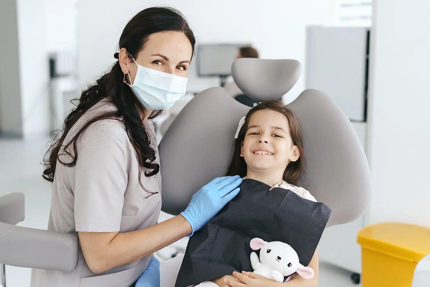 At Levine Orthodontics in Fort Myers, Cape Coral or Lehigh Acres, FL, a children’s orthodontist with a mask and gloves sits beside a smiling young girl holding a stuffed animal in the dental chair.
