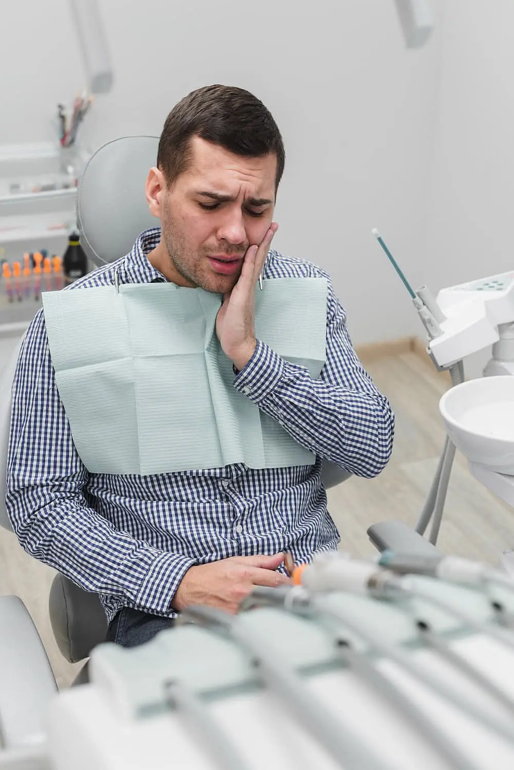 In a dental chair, a man holds his cheek in discomfort, possibly contemplating the Invisalign vs Braces debate, surrounded by dental tools. For those facing similar choices, Levine Orthodontics in Fort Myers, Cape Coral or Lehigh Acres, FL can help.