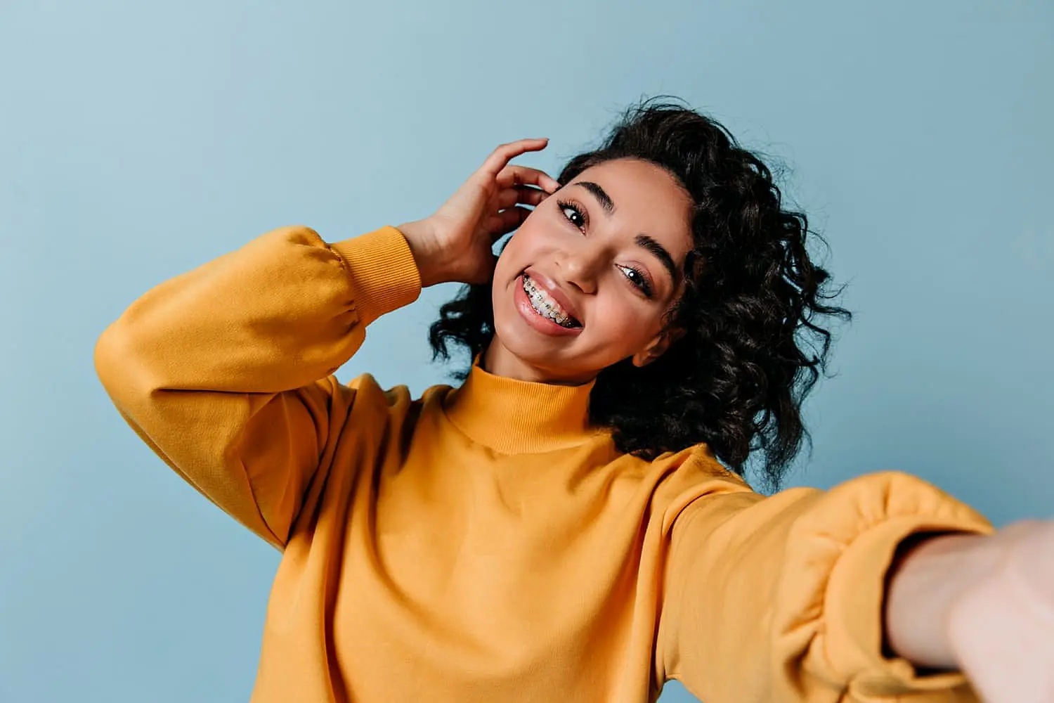 A confident young woman with curly hair in a yellow sweater smiles for a selfie against a light blue background, highlighting the benefits of adult orthodontics at Levine Orthodontics in Fort Myers, Cape Coral or Lehigh Acres, FL.