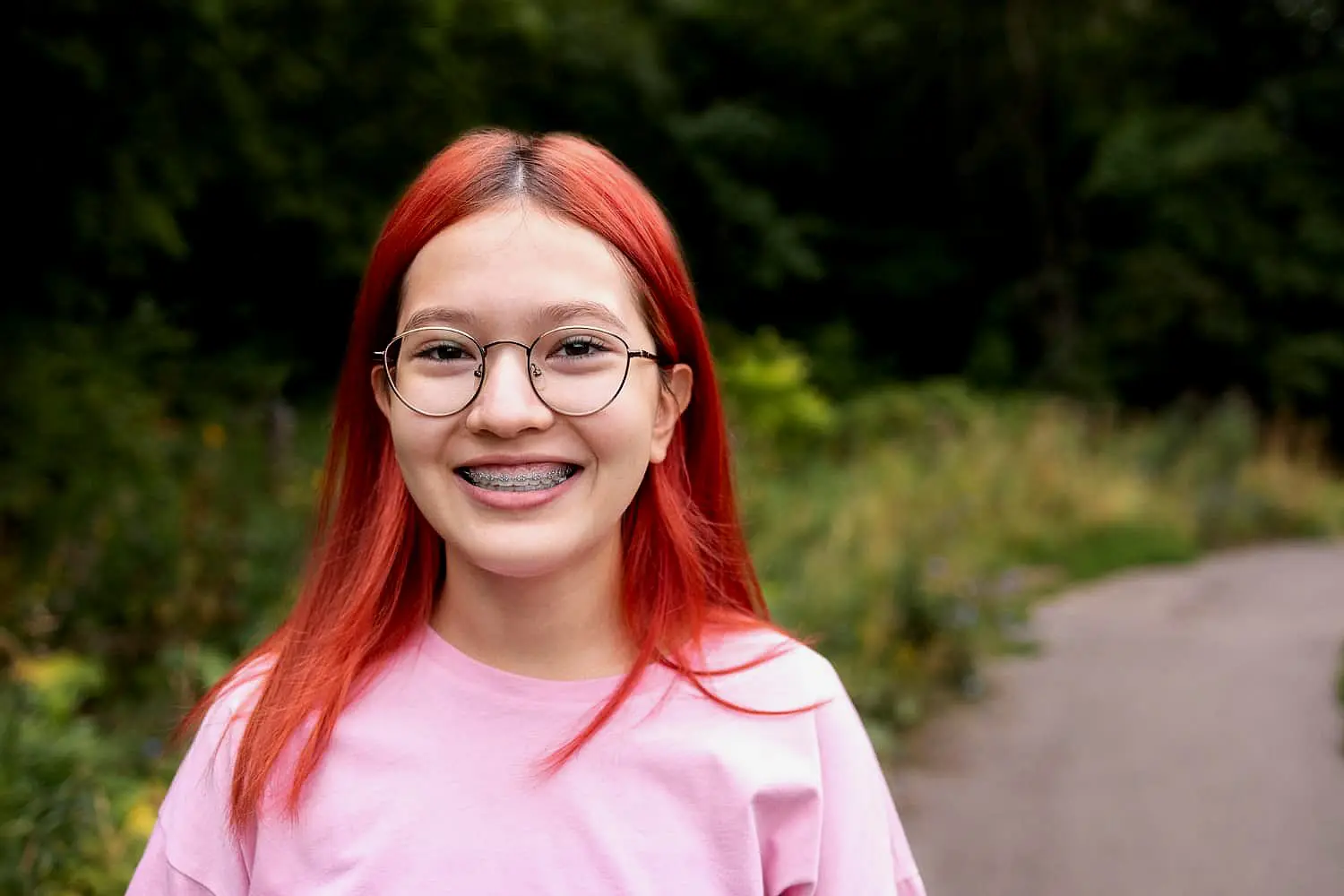 Smiling outdoors on a path surrounded by greenery, a person with red hair, glasses and metal braces showcases the results of Levine Orthodontics in Fort Myers, Cape Coral or Lehigh Acres, FL.