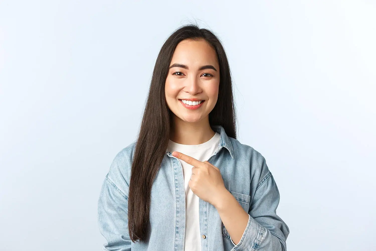 A smiling woman with long dark hair and an overbite, wearing a light denim shirt over a white top, showcases her smile against a plain background. Discover how Levine Orthodontics in Fort Myers, Cape Coral or Lehigh Acres, FL can transform smiles like hers.