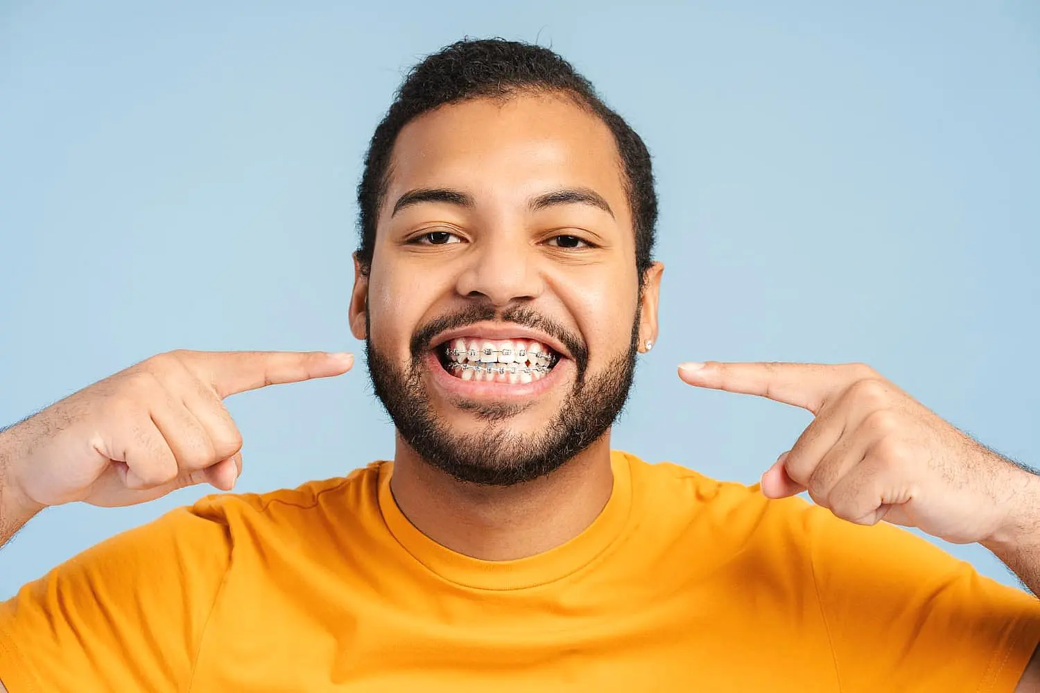 A person in an orange shirt smiles and proudly points to their metal braces with both hands against a light blue background, showcasing the results of Levine Orthodontics in Cape Coral, FL.