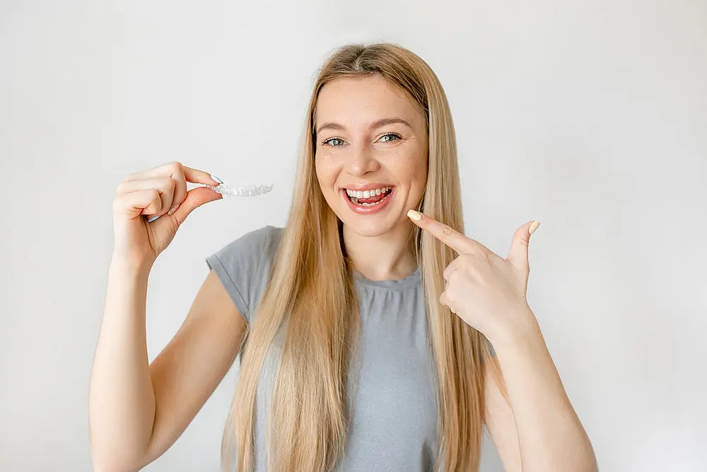 A woman with long blonde hair smiles while pointing to a clear dental aligner, showcasing the benefits of an Invisalign payment plan at Levine Orthodontics in Fort Myers, Cape Coral or Lehigh Acres, FL.