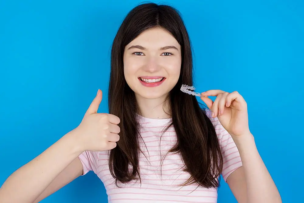 A young woman with long dark hair stands against a solid blue background, smiling confidently and giving a thumbs up while holding a clear dental aligner—implying that Invisalign may be covered by insurance at Levine Orthodontics in Fort Myers, Cape Coral or Lehigh Acres, FL.