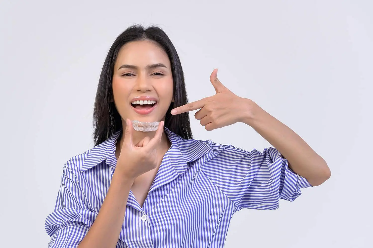 A woman in a striped shirt smiles and points to her mouth while holding Invisalign® Clear Aligners, representing Levine Orthodontics in Fort Myers, FL.