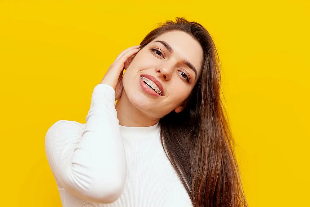 Smiling brightly against a vibrant yellow background, a person with long dark hair and clear braces showcases the quality work of Levine Orthodontics in Fort Myers, FL.