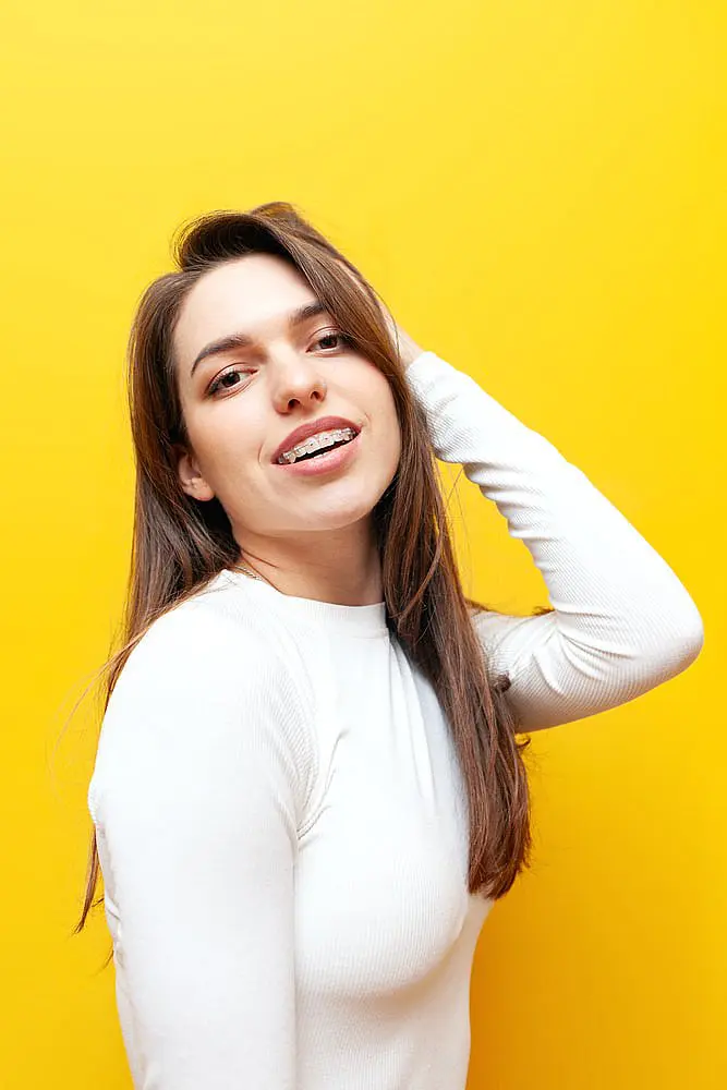 A woman with long brown hair and clear braces, wearing a white long-sleeve top, poses with one hand on her head against a vibrant yellow background—showcasing the results of Levine Orthodontics in Fort Myers, Cape Coral or Lehigh Acres, FL.