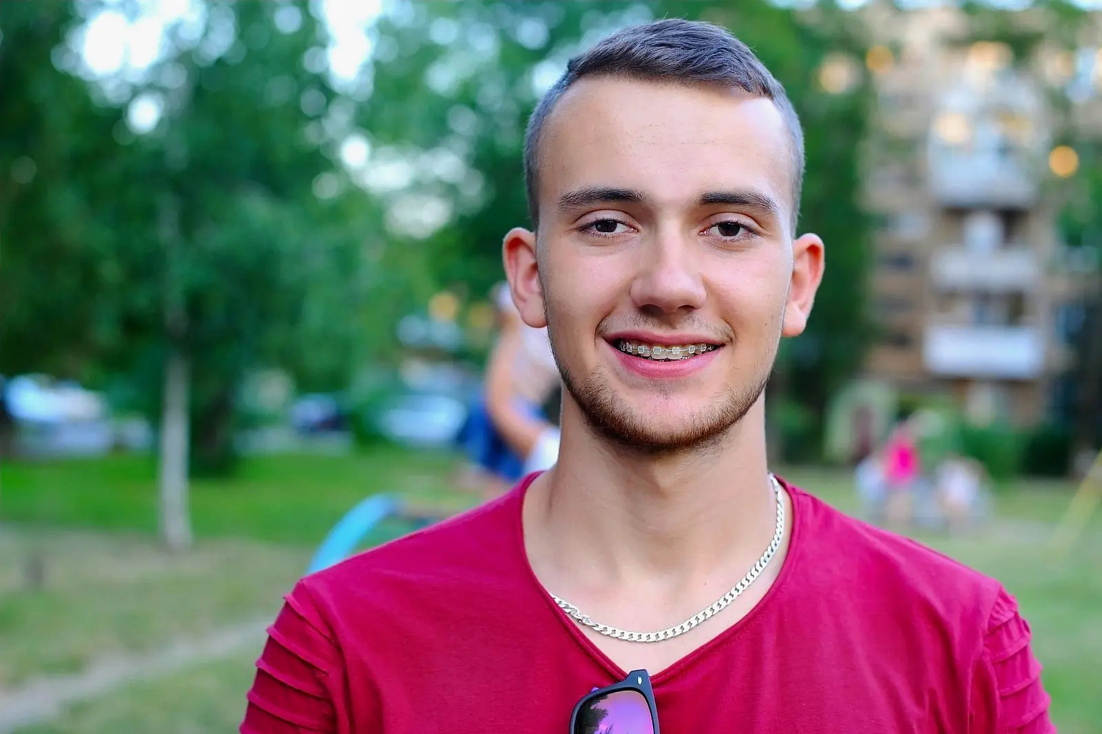A young man sporting short hair, clear braces from Levine Orthodontics in Fort Myers, Cape Coral or Lehigh Acres, FL, a red shirt, and a silver chain stands outdoors in a park with blurred trees and buildings behind him.