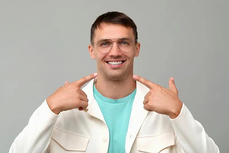 A man in glasses and a cream jacket smiles and points to his adult braces, highlighting the results from Levine Orthodontics in Fort Myers, Cape Coral or Lehigh Acres, FL, against a light gray background.