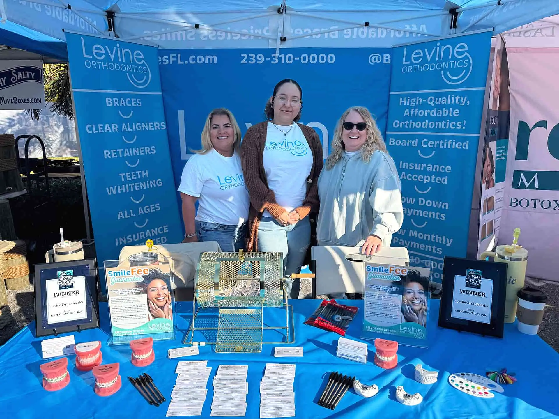 At an outdoor event in Lehigh Acres FL, three women stand behind a blue tablecloth-covered promotional table for Levine Orthodontics, featuring brochures, pens, and dental samples.