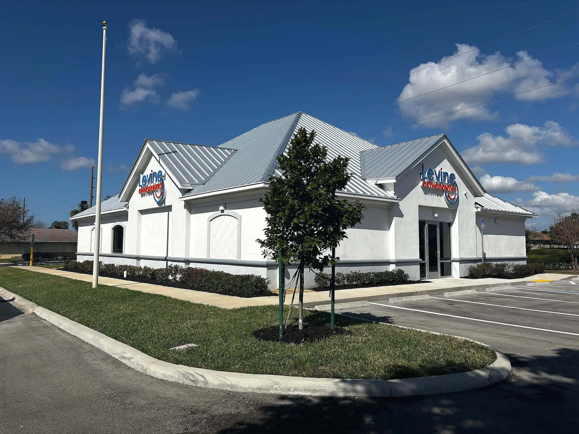 Levine Orthodontics, located in Fort Myers, Cape Coral or Lehigh Acres, FL, operates from a white building with a gray roof. Surrounded by a parking lot and a peaceful grassy area, it offers expert services such as teeth retainer fittings.