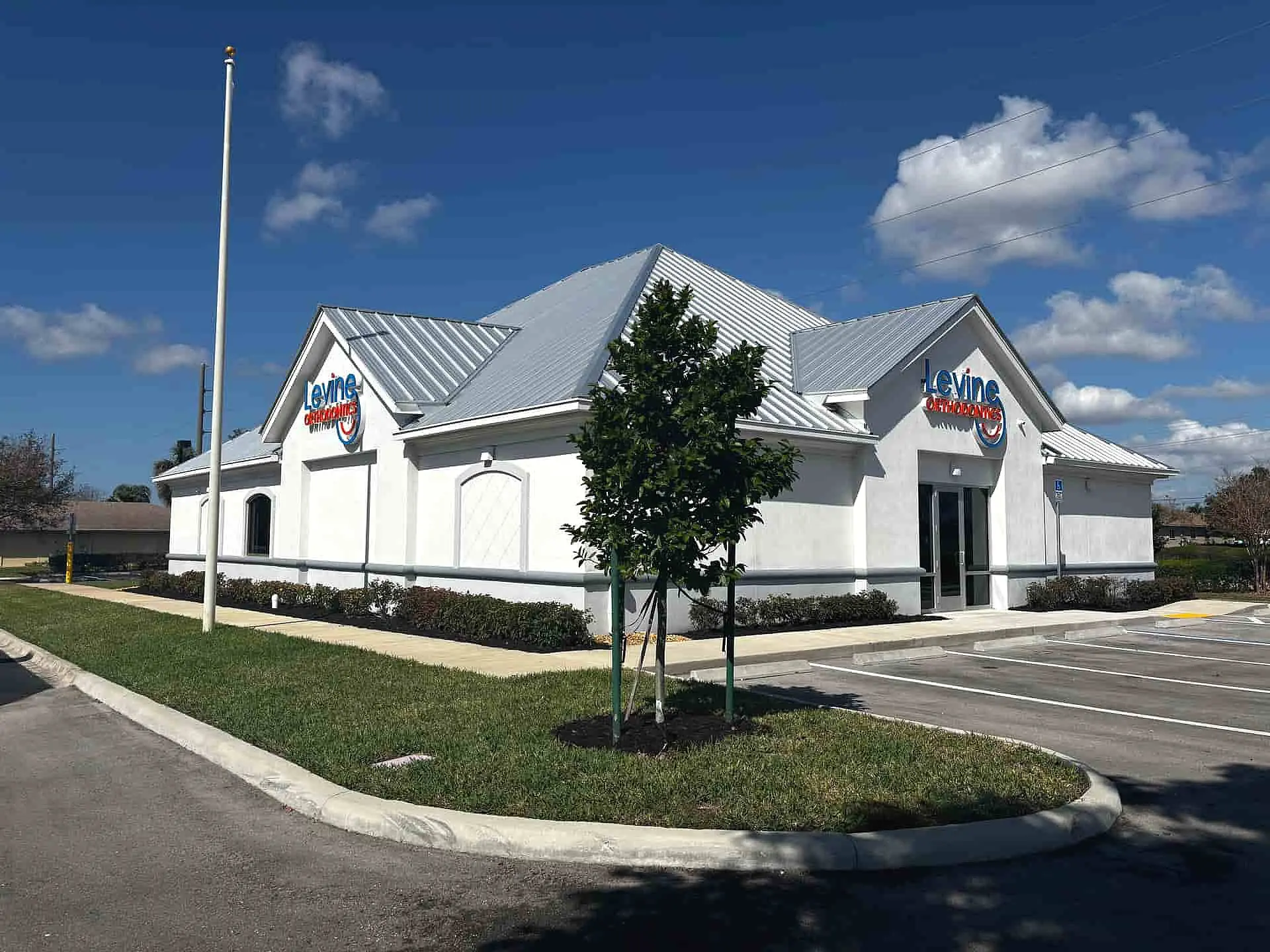 A white single-story building with a metal roof and a "Levine Orthodontics" sign stands beside an empty parking lot under a blue sky with scattered clouds in Cape Coral, FL.