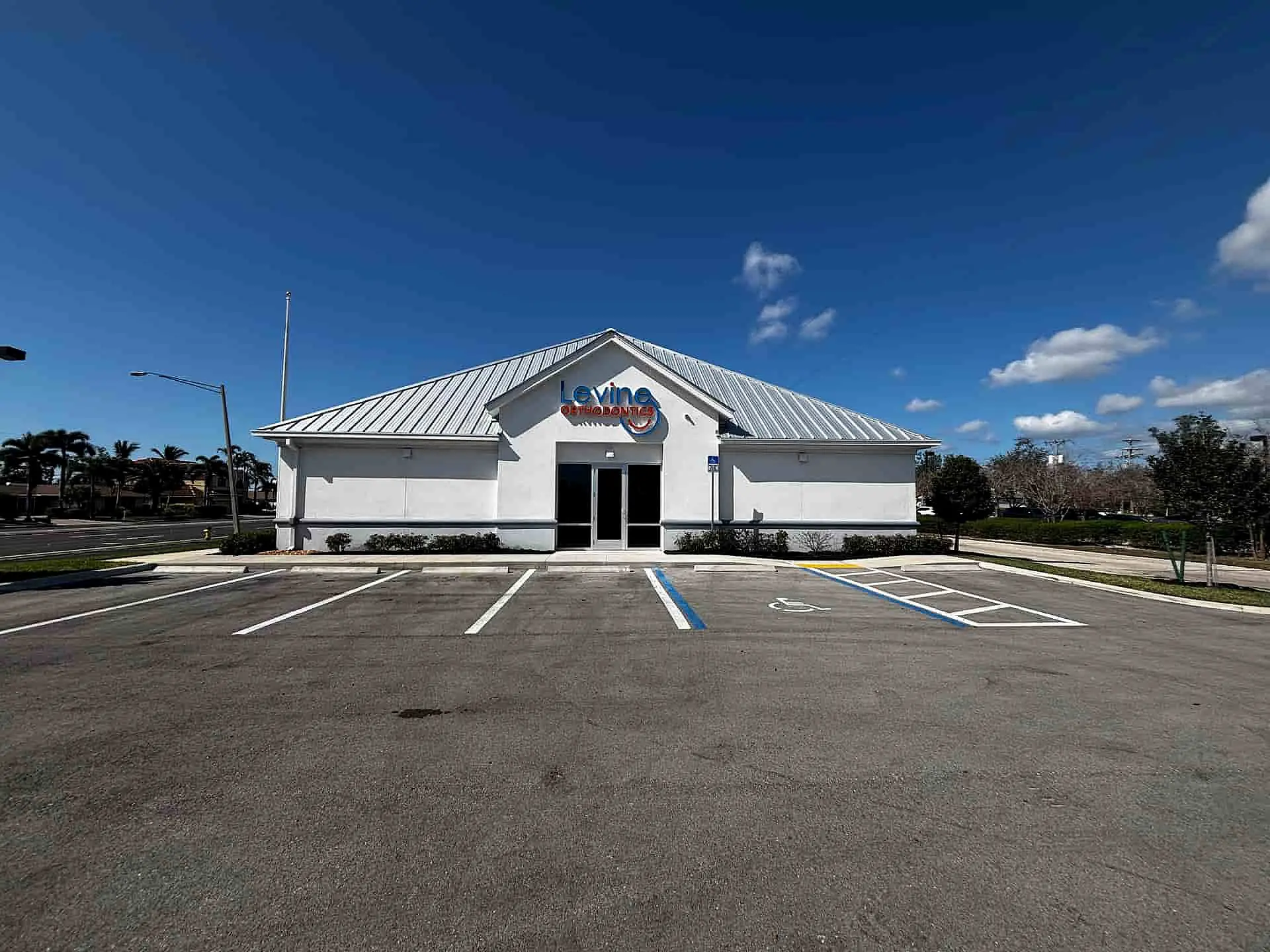 Levine Orthodontics in Fort Myers, Cape Coral or Lehigh Acres, FL, offers affordable Invisalign in a small white single-story building with a metal roof and a "Loving" sign above the entrance, set beside an empty parking lot under a blue sky.