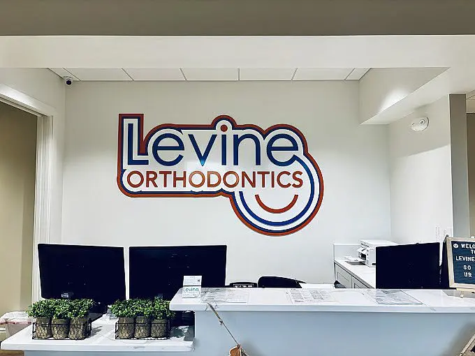 In the reception area of Levine Orthodontics in Cape Coral FL, the logo is displayed on the wall behind a front desk equipped with computers, paperwork and small potted plants.