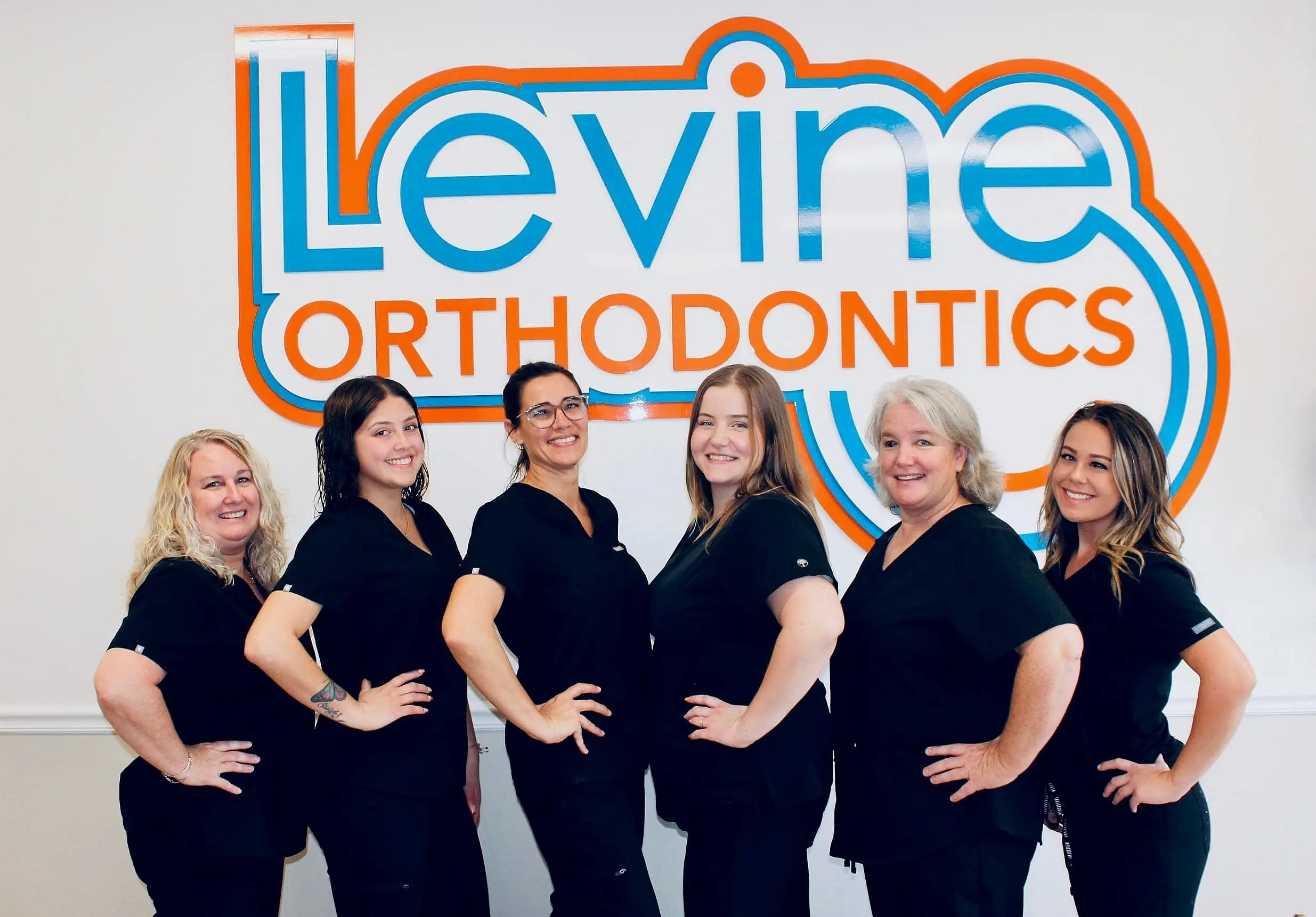 Smiling in front of the Levine Orthodontics sign, six women in black scrubs represent a team of dedicated orthodontic experts serving patients in Fort Myers, Cape Coral, or Lehigh Acres, FL.