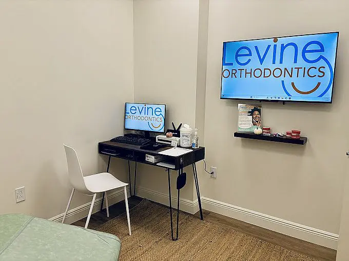 In Fort Myers, Cape Coral or Lehigh Acres, FL, Levine Orthodontics features a small orthodontic office room equipped with a desk, chair, computer, dental supplies and a wall-mounted TV displaying "Levine Orthodontics.
