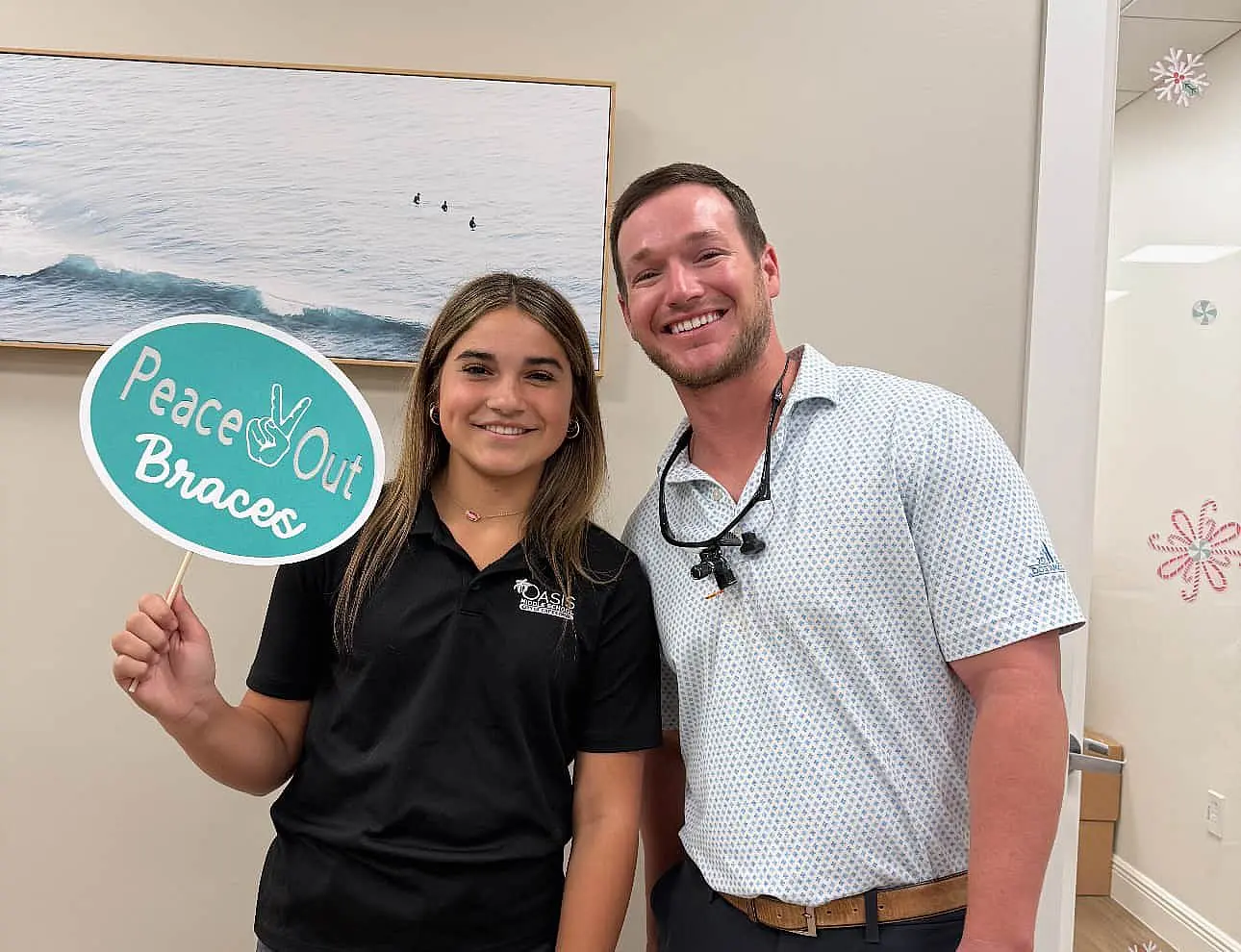 At Levine Orthodontics in Cape Coral FL, two people smile indoors; the person on the left holds a “Peace Out Braces” sign. A framed wave photo hangs on the wall behind them at Dr. Alex Levine’s office.