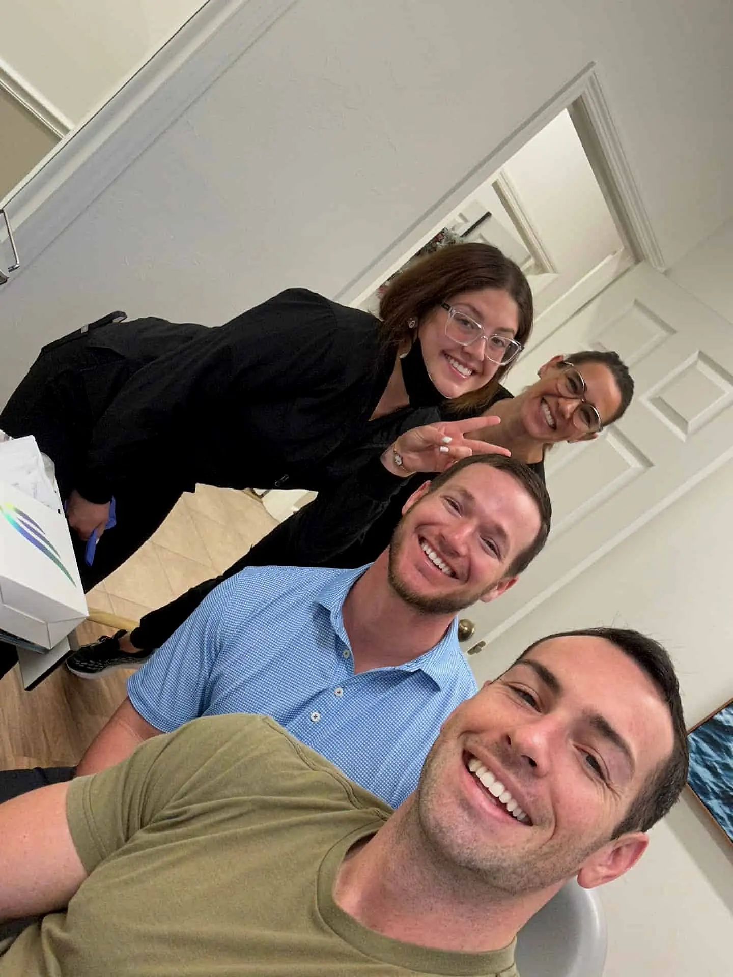 At Levine Orthodontics in Lehigh Acres FL, four adults take a cheerful indoor selfie—two standing behind and two seated in front—while one in back flashes a peace sign to show off their radiant Invisalign smile.