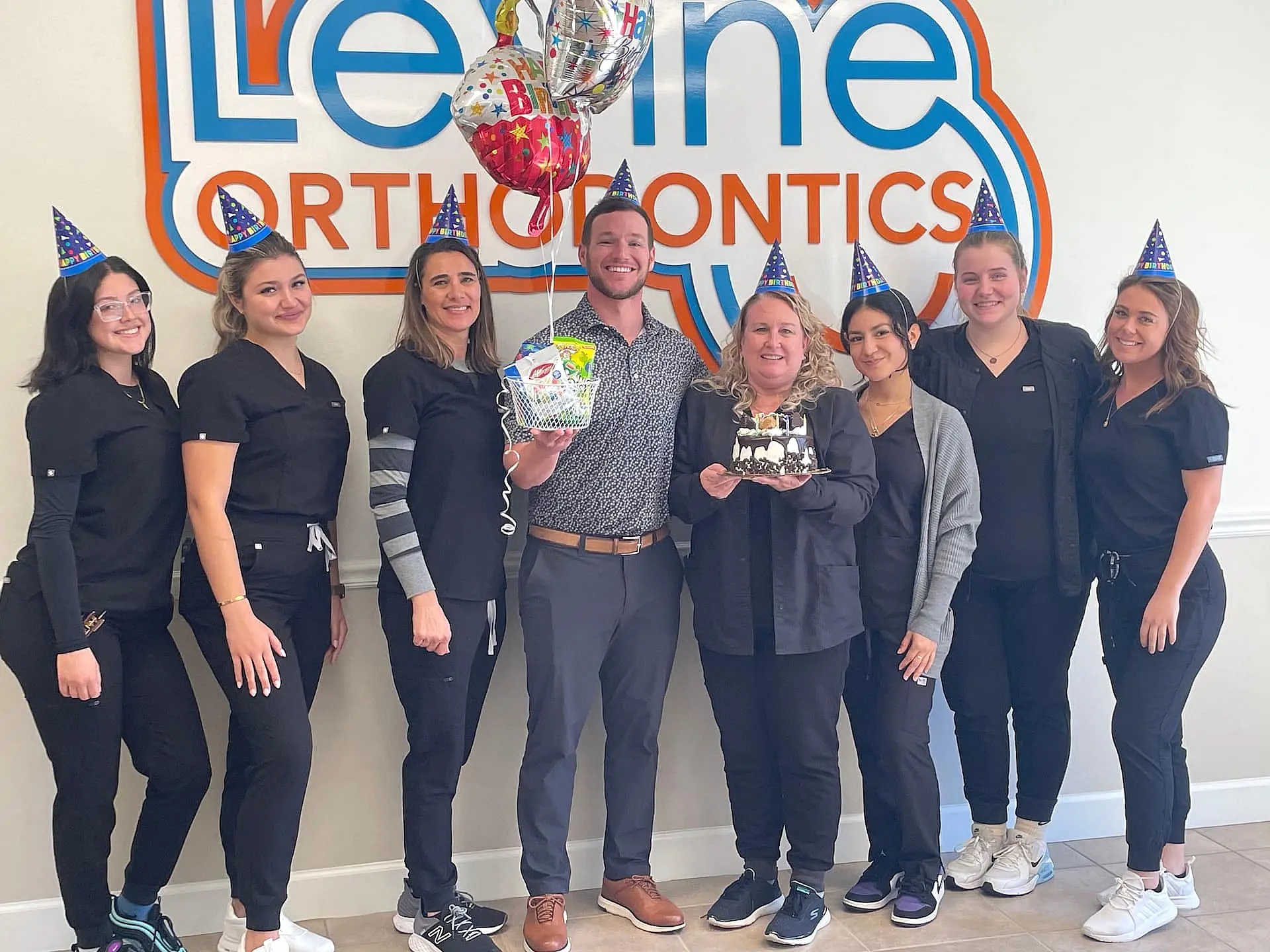 In Fort Myers, Cape Coral or Lehigh Acres, FL, a group of eight in party hats celebrates with Dr. Alex Levine at Levine Orthodontics. They gather before a "Braces and Orthodontics" sign, featuring balloons, a card and a cake—ideal for any mouth breather's sweet tooth.