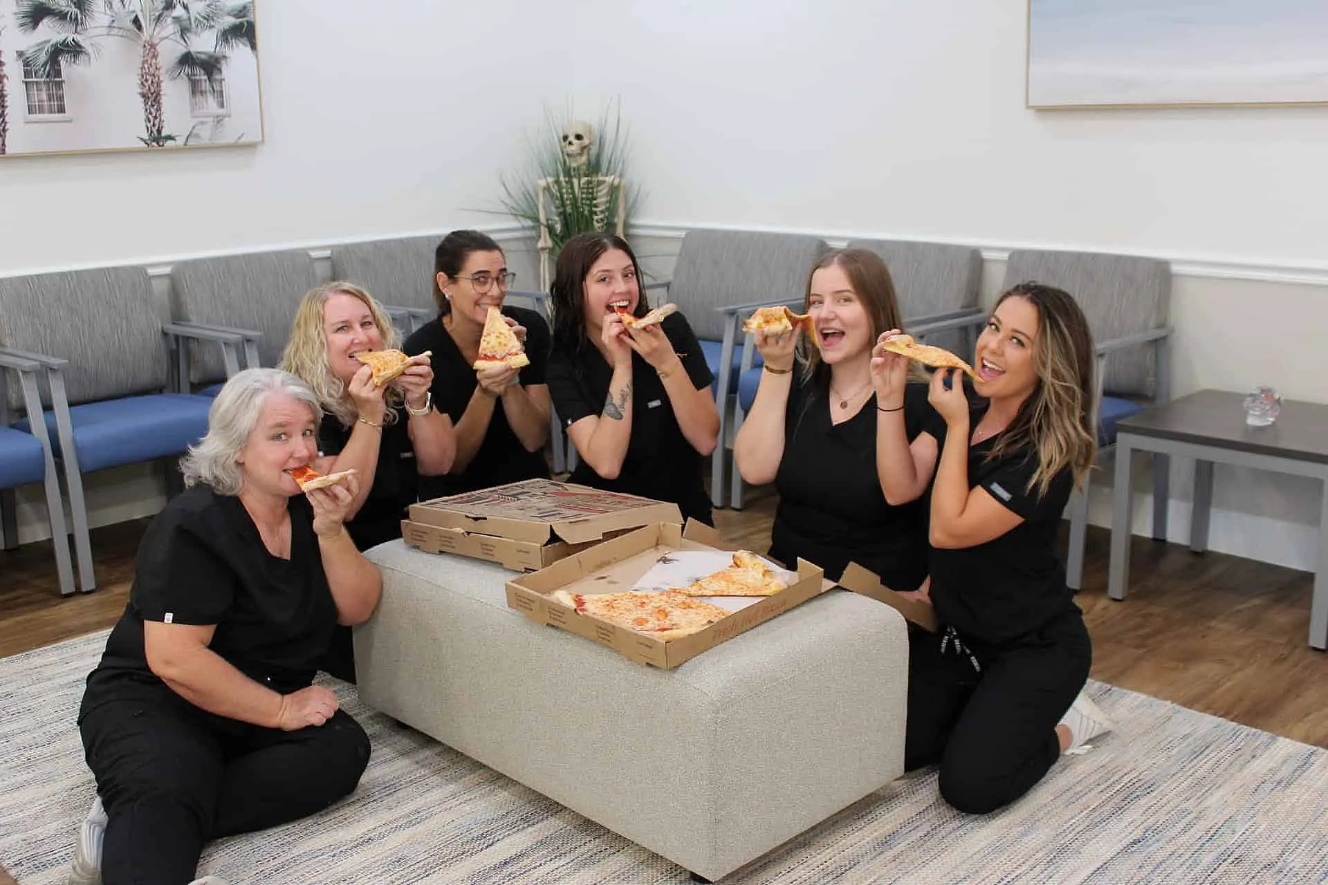 At Levine Orthodontics in Fort Myers, Cape Coral or Lehigh Acres, FL, six women in black uniforms enjoy pizza together around a table in the waiting room, smiling for the camera.