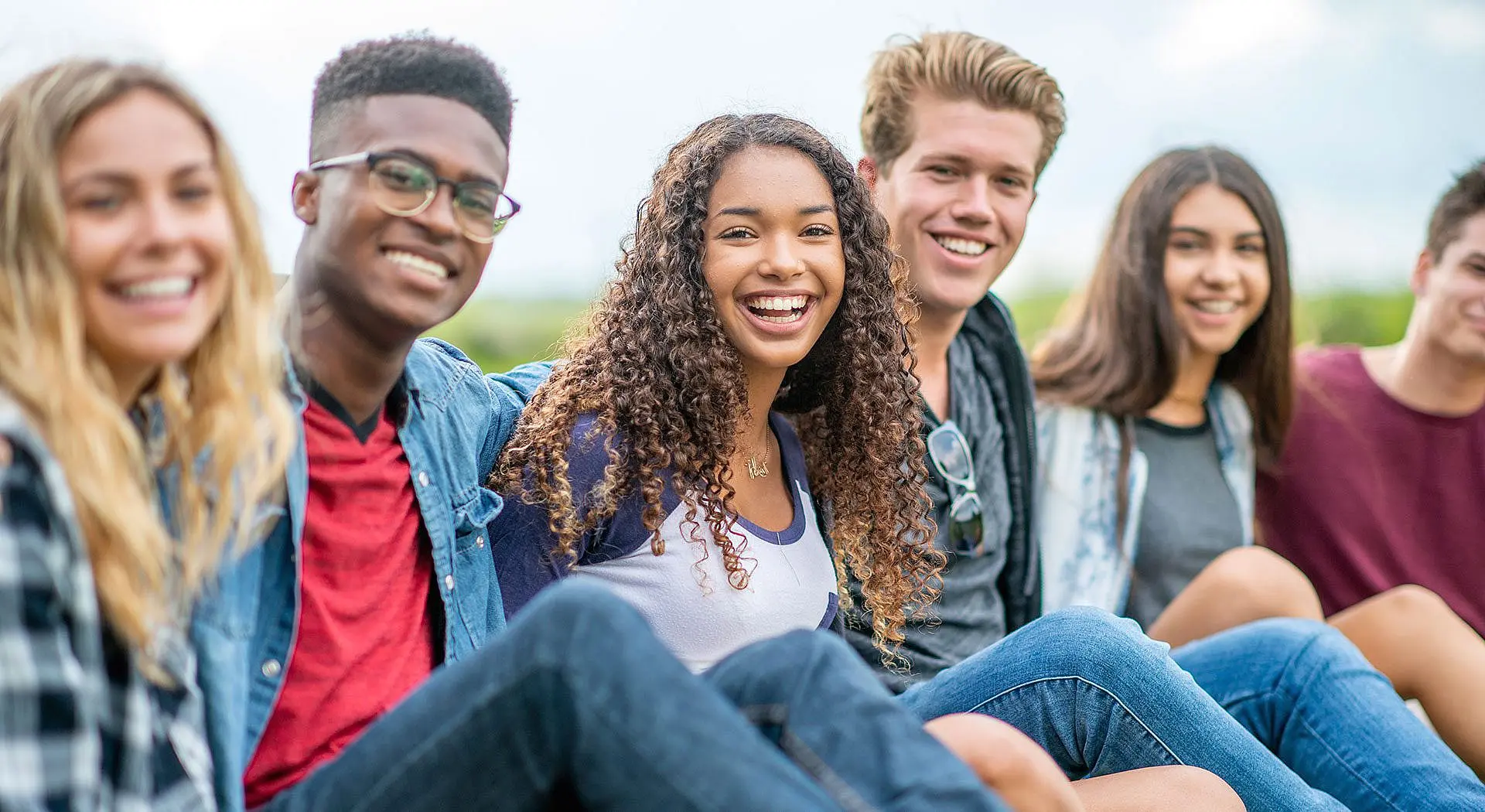 A group of six young people sit outdoors in a row, smiling at the camera—some proudly displaying their braces from Levine Orthodontics in Fort Myers, FL.
