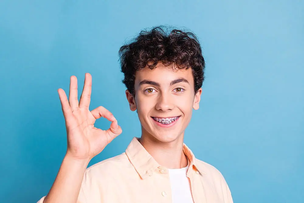 A teen boy with curly hair and braces stands in front of a blue background, smiling brightly and giving an "OK" hand gesture—representing the confident results achieved at Levine Orthodontics in Cape Coral, FL.