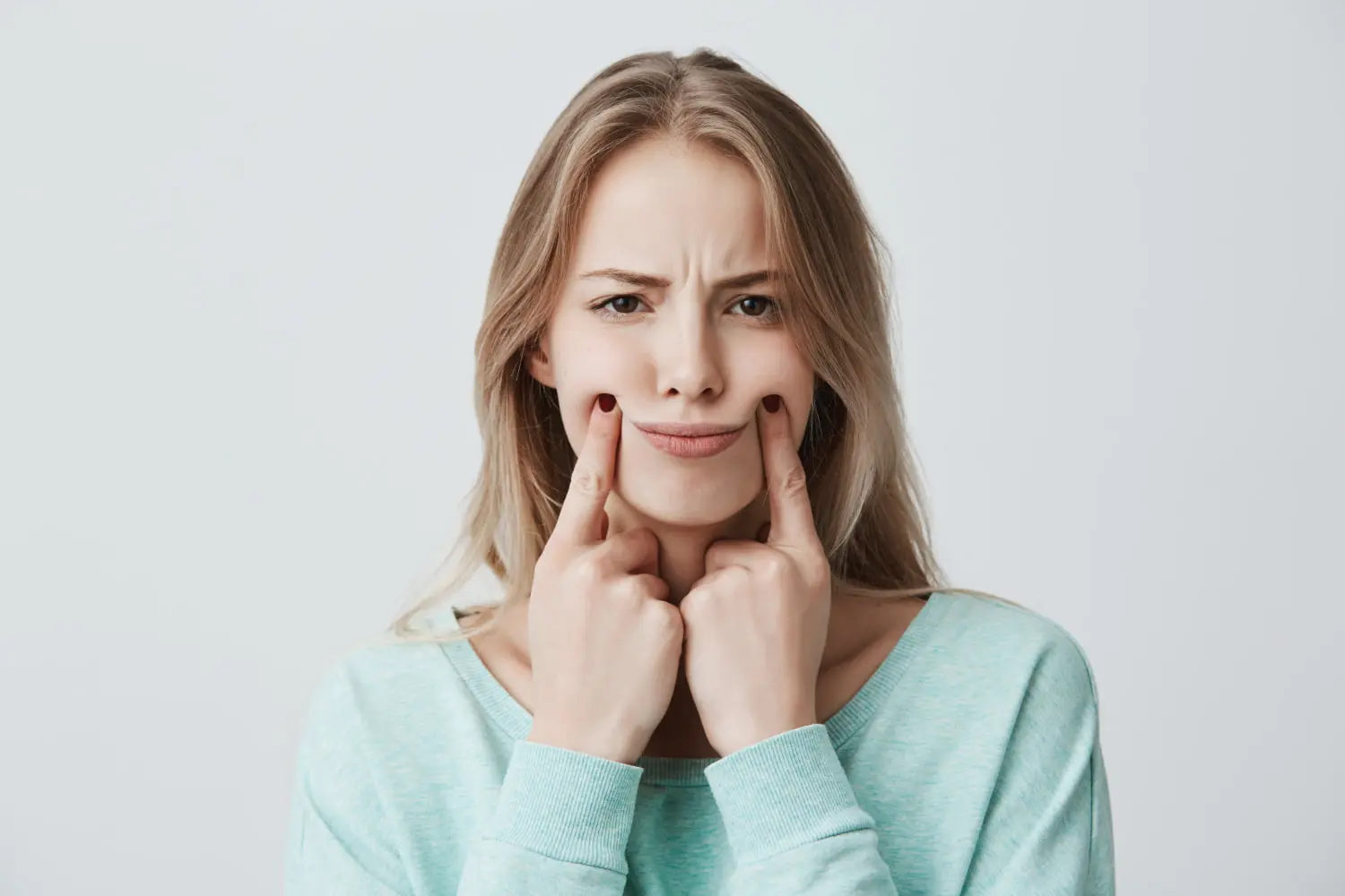 At Levine Orthodontics in Fort Myers, Cape Coral, or Lehigh Acres, FL, a young woman with long blonde hair wears a light blue top and presses her fingers to the corners of her mouth for a forced smile that reveals a slight overbite against a plain background.